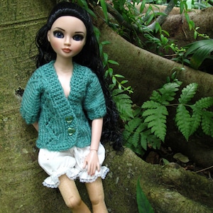 Peut inclure: Une poupée aux longs cheveux noirs portant un cardigan en tricot turquoise et une culotte en dentelle blanche. La poupée est debout devant un tronc d'arbre avec un feuillage vert.