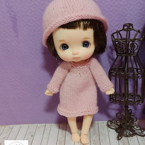 May include: A small pink knitted doll wearing a pink knitted dress and a pink knitted hat. The doll is standing on a wooden surface.