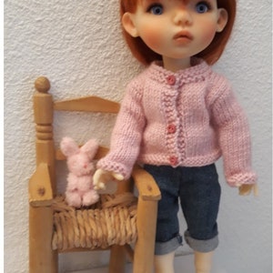 Peut inclure: Une poupée aux cheveux roux et aux yeux bleus portant un cardigan rose tricoté et un jean bleu. Un petit lapin rose est assis sur une chaise en bois miniature. Le texte "Ma poupée tricote" est visible.