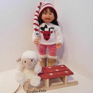 May include: A doll wearing a red and white striped hat, a red and white striped sweater with a reindeer design, and red and white striped pants. The doll is holding a silver bell and is sitting on a red wooden sled with white snowflakes painted on it. A white stuffed sheep is sitting on the sled.