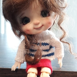 May include: A smiling doll with brown curly hair wearing a white and blue striped sweater and a red skirt. The doll has large green eyes and rosy cheeks.
