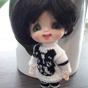 May include: A small doll with black hair and green eyes wearing a white and black knitted sweater and striped pants. The doll is smiling and the text "Ma poupée tricote" is visible in the background.
