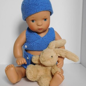 以下が含まれることがあります： 白い表面に座っている人形。青いニット帽、青いニットベスト、それに合わせたおむつを着用し、茶色のぬいぐるみウサギを抱えています。