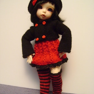 May include: A black knitted doll with red and black striped stockings, wearing a black knitted dress with a red knitted bodice and a black hat with a red bow.