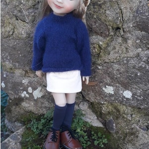 Può includere: Una bambola con i capelli biondi che indossa un maglione blu, una gonna bianca e scarpe marroni. La bambola è in piedi davanti a un muro di pietra.