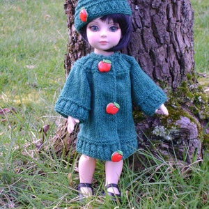 Könnte beinhalten: Ein grünes Strickkleid für Puppen mit roten Apfelknöpfen. Die Puppe trägt eine passende grüne Strickmütze. Die Puppe steht vor einem Baumstamm.