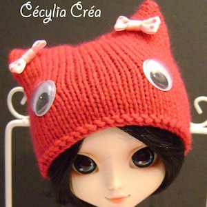 May include: A red knitted hat with cat ears and pink bows. The hat has large, black googly eyes. The hat is worn by a doll with black hair and large, blue eyes.