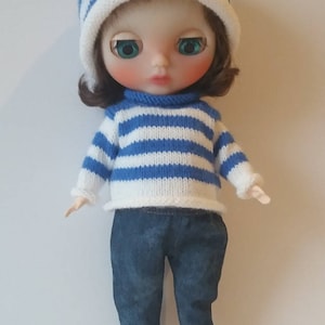 May include: A doll wearing a blue and white striped sweater, blue jeans with plaid trim, and a blue and white striped hat.