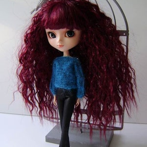 May include: A doll with long, curly, burgundy hair, wearing a blue sweater and black pants. The doll is standing in front of a gray birdcage.