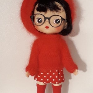May include: A red-haired doll wearing a red jumper, a red and white polka dot skirt, red socks, and black shoes. The doll is wearing black framed glasses and has red lipstick. The doll is standing on a white surface. The text "Ma poupée tricote" is written in black at the top of the image.