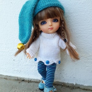 Peut inclure: Une petite poupée portant un pull en laine blanc, un legging rayé bleu et blanc et un bonnet bleu en laine avec une clochette dorée. La poupée a de longs cheveux bruns et des yeux bleus.