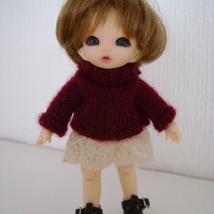 May include: A small doll with brown hair, blue eyes, and a burgundy sweater. The doll is wearing a white lace skirt and black shoes.
