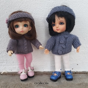 以下が含まれることがあります： 手編みの衣装を着た2体の人形。1体はグレーのセーター、ピンクのレギンス、ピンクの靴を履いています。もう1体はグレーのセーター、白いレギンス、青い靴を履いています。どちらの人形も青い目と黒髪です。「Cécylia Créa」の文字が下にあります。