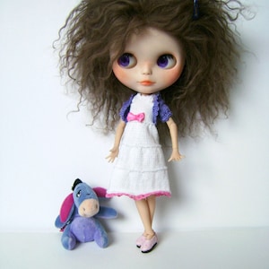 May include: A doll with long brown curly hair wearing a white dress with a purple cardigan and pink shoes. The doll is holding a purple stuffed animal.