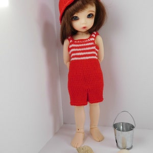 May include: A red and white striped knitted swimsuit with a red knitted hat on a doll with brown hair and blue eyes. The doll is standing on a white surface with seashells and a small metal bucket.