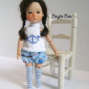 May include: A doll with brown hair in braids wearing a white knitted top with a blue design, blue and white striped knee-high socks, and blue and white patterned shorts. The doll is standing next to a white wooden chair with a woven seat.