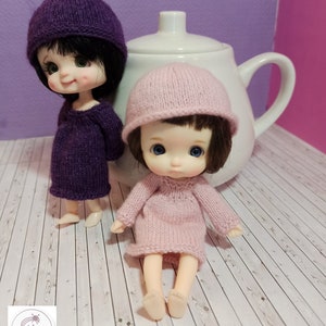 May include: Two small dolls wearing knitted hats and dresses. The doll on the left is wearing a purple hat and dress. The doll on the right is wearing a pink hat and dress. The dolls are sitting on a white wooden surface.
