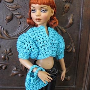 Peut inclure: Une poupée portant un haut en crochet bleu et un sac en crochet bleu. La poupée a les cheveux roux et porte un pantalon noir.