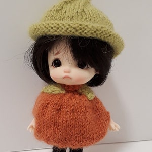 May include: A handmade doll wearing a knitted outfit. The doll has dark hair and is dressed in an orange sweater, black pants, and a green hat. The text "Ma poupée tricote" is at the top of the image.
