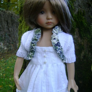 May include: A white dress with a lace trim and a white knitted cardigan with a floral print. The doll has brown hair and blue eyes.