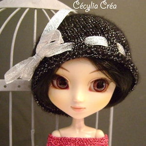 May include: A black knitted hat with silver glitter and a silver ribbon bow. The hat is worn by a doll with black hair and large, bright eyes.