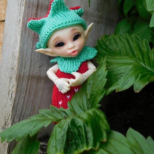 Puede incluir: Una pequeña muñeca vestida con un traje de fresa de punto rojo y verde. La muñeca tiene grandes ojos azules y está posada con las manos juntas. La muñeca está sentada frente a un fondo verde frondoso.