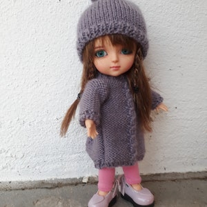 Könnte beinhalten: Eine kleine Puppe, die einen lila gestrickten Hut mit Bommel, einen lila gestrickten Mantel und rosa Leggings trägt. Die Puppe trägt lila Schuhe und hat lange braune Haare.