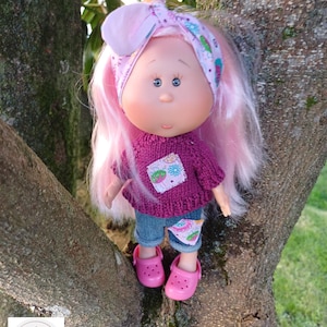 May include: A doll with long pink hair wearing a purple knitted sweater with a floral patch, blue denim cuffed pants with a floral patch, and pink clogs. The doll has a pink floral headband tied in a knot on top of its head.