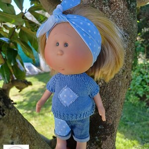 Könnte beinhalten: Eine Puppe mit blondem Haar trägt ein hellblaues, gestricktes Kurzarmshirt mit einem weiß gepunkteten Aufnäher, eine blaue Jeansshorts mit einem passenden gepunkteten Aufnäher und ein hellblaues Stirnband mit weißen Punkten, das oben gebunden ist.