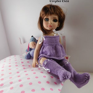 Può includere: Un vestito di maglia viola e leggings abbinati con dettagli di nastro bianco su una bambola con capelli castani e occhi azzurri. La bambola è seduta su un letto bianco con pois rosa.