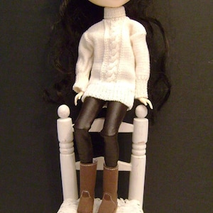 May include: A doll with black hair and blue eyes is sitting on a white wooden chair. The doll is wearing a white sweater and brown boots. The chair has a woven seat.