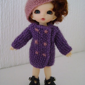 May include: A small doll with brown hair wearing a purple knitted coat with pink stars, a pink knitted hat, and black shoes.