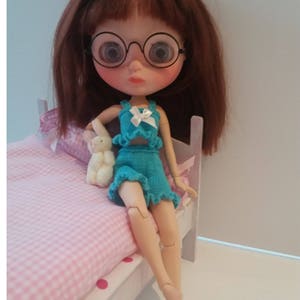 May include: A doll wearing a turquoise knitted outfit with a white bow, sitting on a pink and white checkered bed with a white teddy bear.