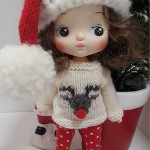 以下が含まれることがあります： 白いニットのセーターにトナカイのデザイン、赤と白のドット柄のレギンス、白いポンポンが付いた赤いニットのサンタ帽をかぶった小さな人形です。