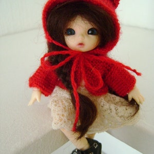 May include: A small doll with long brown hair wearing a red knitted hooded sweater and a white lace skirt. The doll is wearing black shoes with buckles.
