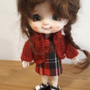 May include: A small doll with brown hair wearing a red knitted cardigan and a red and black plaid dress. The doll is wearing black shoes with white cat faces on the toes.