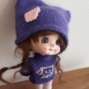 May include: A purple knitted hat with a pink button on top, worn by a doll with brown hair, green eyes, and a purple knitted sweater with a white design. The doll is smiling and has rosy cheeks.