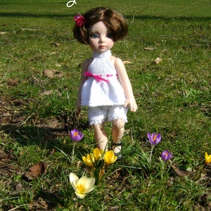 May include: A porcelain doll with brown hair wearing a white dress with a pink sash and a pink flower in her hair. The doll is standing in a grassy field with yellow and purple crocuses.