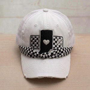 May include: A white distressed baseball cap featuring a central black patch shaped like the state of Indiana with a white heart. The patch is flanked by black and white checkered panels with silver studs and a black and white checkered fabric bow.