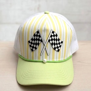 May include: A trucker hat featuring a yellow and white vertical striped front panel with an embroidered patch of two crossed black and white checkered racing flags. The hat has a light green brim, a green cord trim, and a white mesh back panel.