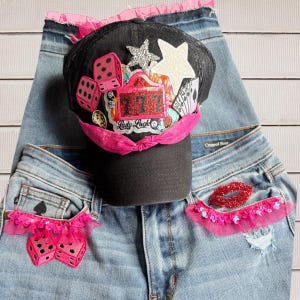 May include: A black baseball cap featuring a pink fabric band and patches of dice, stars, playing cards, and a red 777 slot machine graphic with the text Lady Luck, resting on light blue denim jeans with pink sequined ruffles and a red lip patch.
