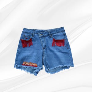 May include: Light blue denim shorts with red fringe pocket accents and a frayed hem. The shorts feature an embroidered flame design at the bottom. A stylish and unique addition to any wardrobe.