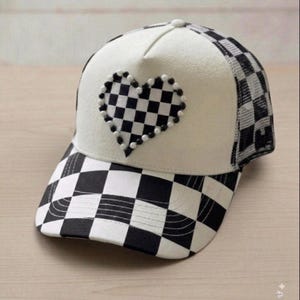 May include: A baseball cap featuring a white front panel with a black and white checkered heart applique outlined in small beads. The brim and side panels display a black and white checkered pattern, while the back consists of mesh fabric.