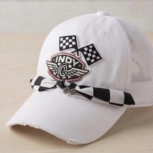 May include: A white baseball cap with a mesh back and a distressed brim. The front features an embroidered patch with the text INDY, a winged wheel, and two checkered flags. A black and white checkered fabric ribbon is tied around the base of the crown.