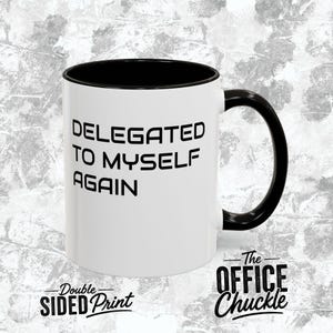 May include: A white ceramic mug with a black interior and a black handle. The exterior features the black text DELEGATED TO MYSELF AGAIN in a bold sans-serif font. The mug is positioned against a textured gray background with branding text.