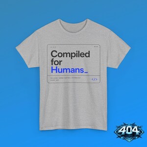 May include: A heather gray short-sleeve t-shirt featuring a black and blue rectangular graphic with the text Compiled for Humans, Building human-centric technology every day, and a code bracket symbol, centered on the chest area.