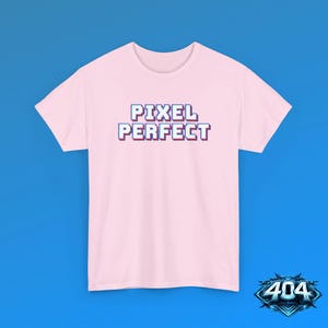 May include: A light pink short-sleeve t-shirt featuring the text PIXEL PERFECT in a blue and white blocky, pixelated font style centered on the chest. The garment has a crew neckline and a standard fit, showcasing a clean and simple design.