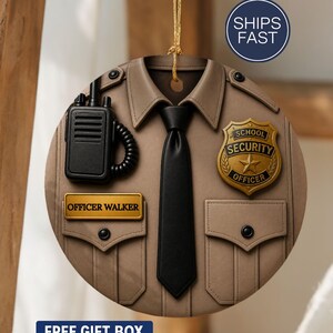 May include: A round ornament resembling a school security officer's uniform. The tan-colored ornament features a black tie, a radio, and a badge that reads "School Security Officer." The name "Officer Walker" is displayed on a name tag. The ornament hangs from a gold string.
