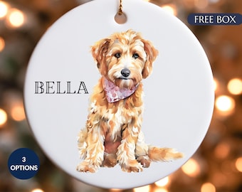 May include: A round white ceramic ornament featuring a painterly illustration of a golden-colored dog wearing a pink patterned bandana. The name BELLA is printed in black capital letters to the left of the dog. A small hole is at the top.