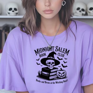 May include: A lilac-colored t-shirt featuring a graphic design for the "Midnight Salem Book Club." The design includes a skull wearing a witch's hat, bats, a pumpkin, and the text "Books and Brews at the Witching Hour!"